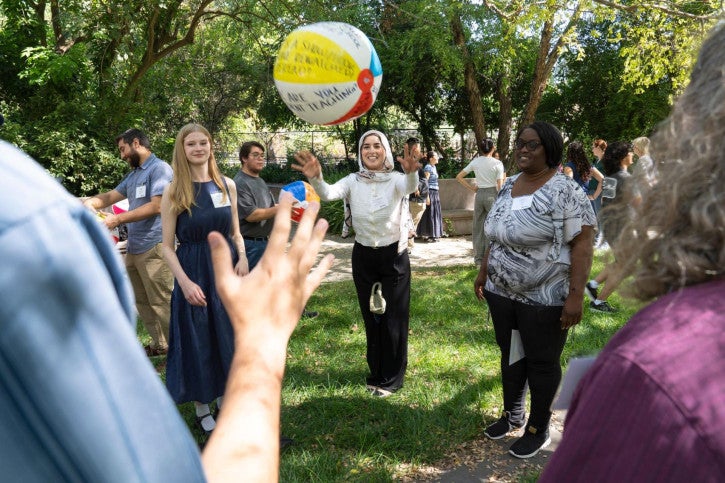 A small group of people stand in a circle outside and take turns throwing a beach ball to each other as an icebreaker activity.