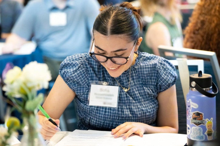 A credential student wearing a blue shirt, glasses, and a nametag sits at a table and writes in her program handbook.