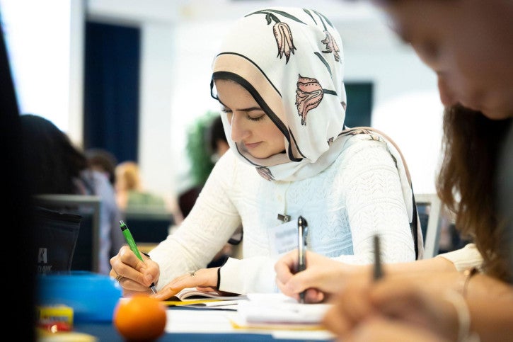 Two credential students sit at a table and look down while writing in their handbooks.