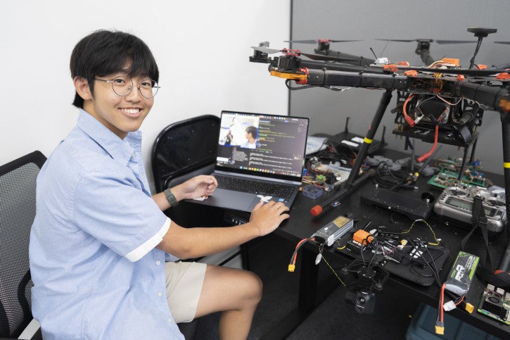 Jayden smiles for the camera while sitting at a laptop. In the background, a drone is sitting on the desk.