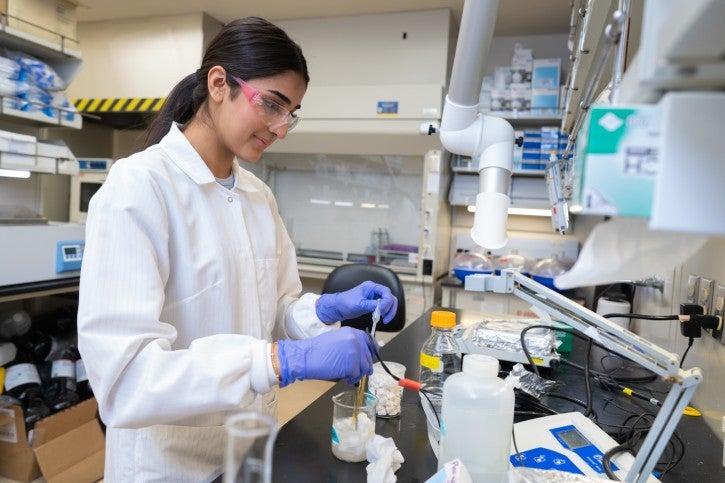 Student wearing gloves and white lab coat works at lab station