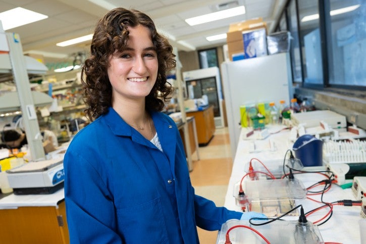 Student wearing gloves and blue lab coat works at lab station
