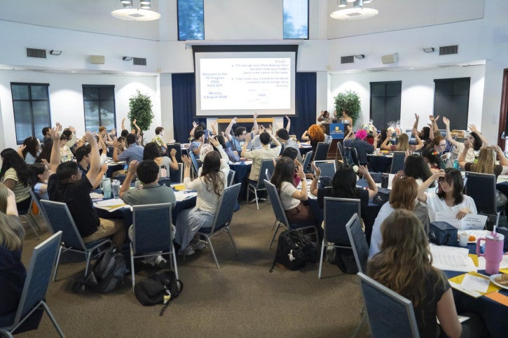 A crowded room of new credential students. They face a projection screen and podium.