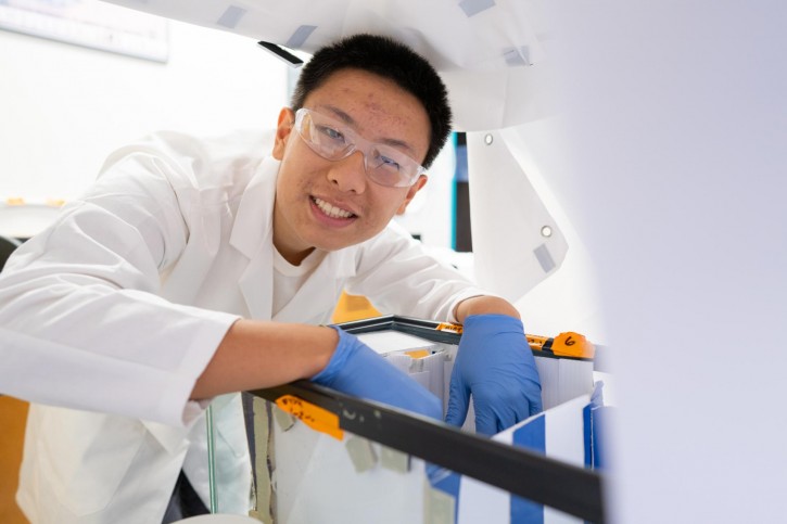 Kyle smiles for the camera, wearing safety goggles, a lab coat, and blue gloves