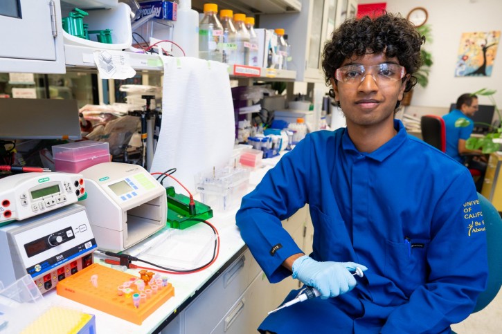 Pranav smiles for the camera, wearing a blue lab coat and holding a syringe