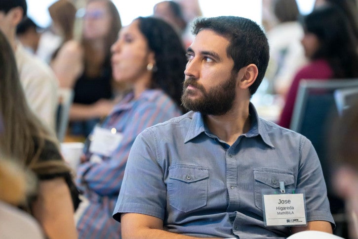 A credential student wearing a blue button-up shirt and nametag looks to the speaker at the front of the room.
