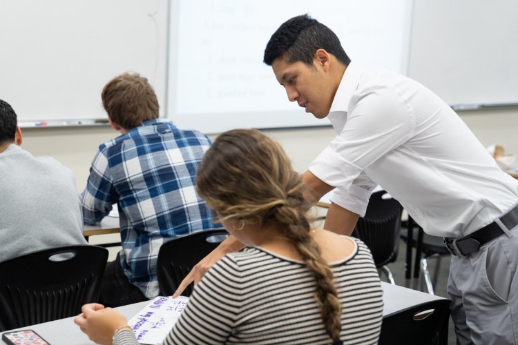 A male teacher wearing a white shirt stands next to a student to explain a math problem.