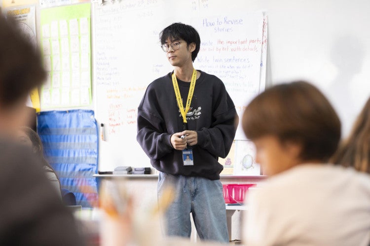 A teacher stands at the front of the classroom and looks off to the side. His students are blurred in the foreground.