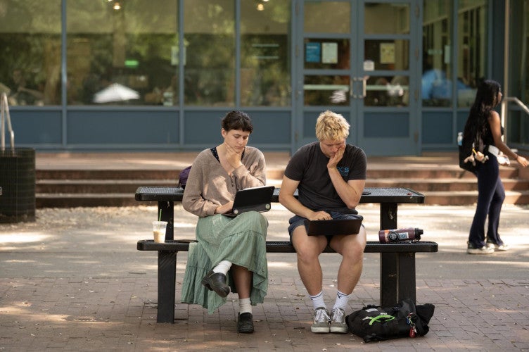 Two students sit at a picnic table outside. They both look down at their laptops in confusion.