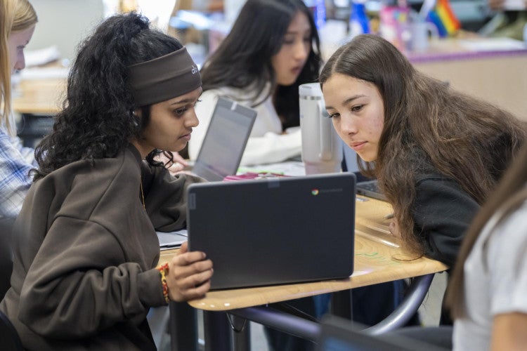 Two students sit at a desk cluster and look at a laptop screen with intensity.