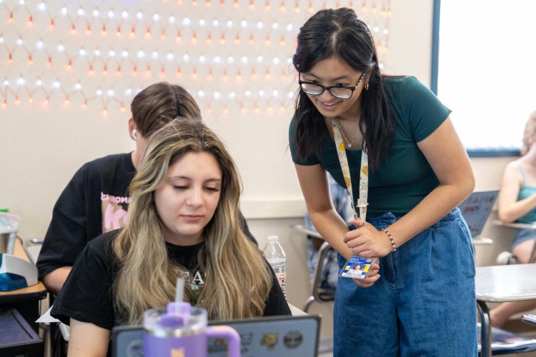 A teacher wearing a lanyard and glasses leans over a student's desk. They both look at a laptop screen together.