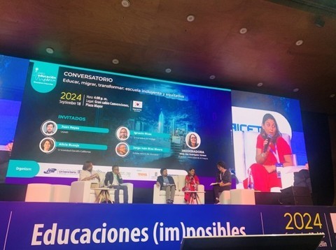 A presentation screen shows four people sitting on a stage, while  one person speaks into a microphone