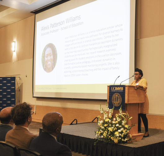 Alexis Patterson Williams stands at a podium and addresses an audience. On a presentation screen behind her is her name, headshot, and biography.