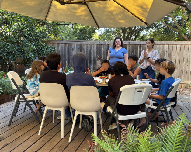 Two women stand next to an outdoor table, shaded by a large umbrella. Seated at the table are a group of elementary school students.