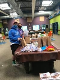 Two men wearing sweatshirts and face masks stand at a table in a warehouse, putting at-home maker kit supplies into paper grocery bags.