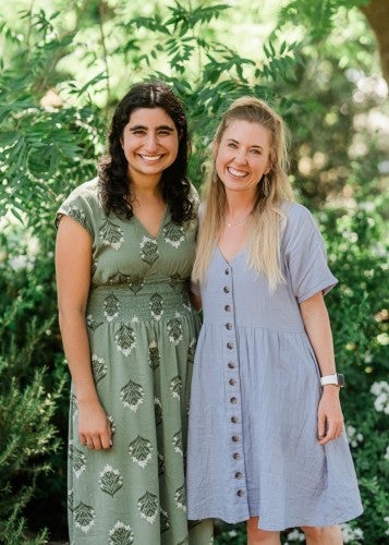 Two women wearing dresses stand arm in arm, posing for a photo.