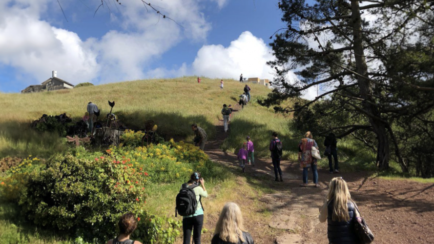 A line of people wearing backpacks walk up a grassy hill with blue skies and clouds in the background.