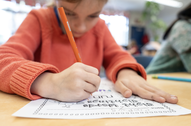 Close up of a young student wearing an orange shirt and writing on a worksheet with a pencil.