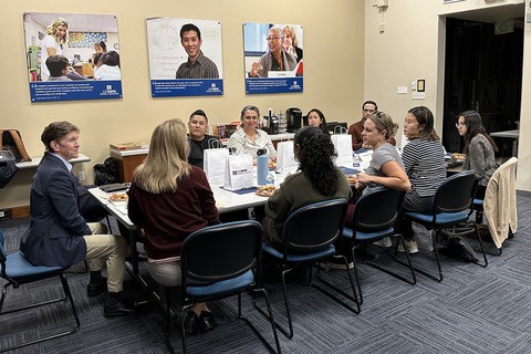 A group of 10 people, sit at a table, facing each other and talking.