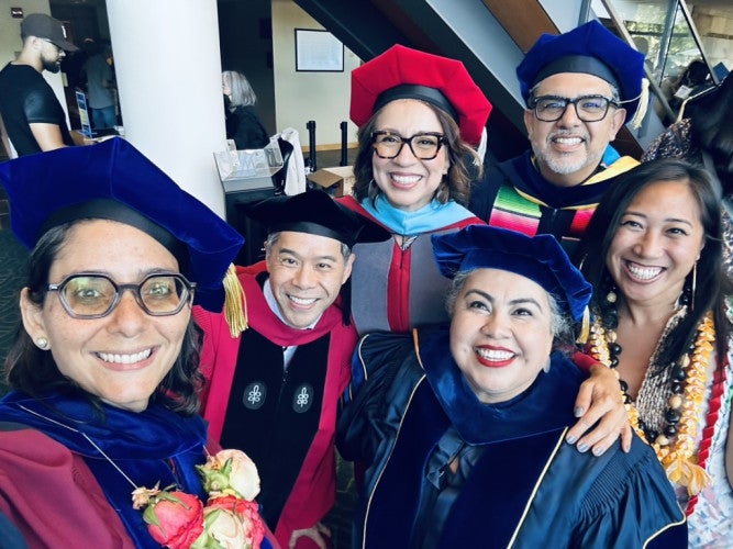 A group of people wearing doctoral graduation regalia pose for a group photo in the Mondavi Center for the Arts.