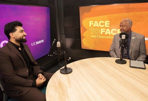 Two men wearing suits sit at a table and face each other.