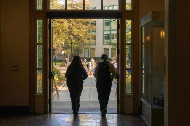 The silhouettes of two students as they step out of a building and onto the UC Davis campus.