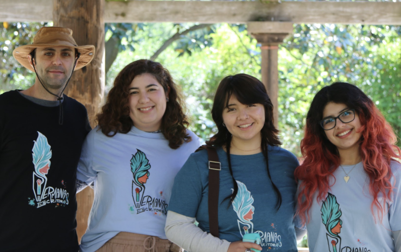 A group of four graduate students stand arm in arm. Their shirts read "Hermanas Escritoras."