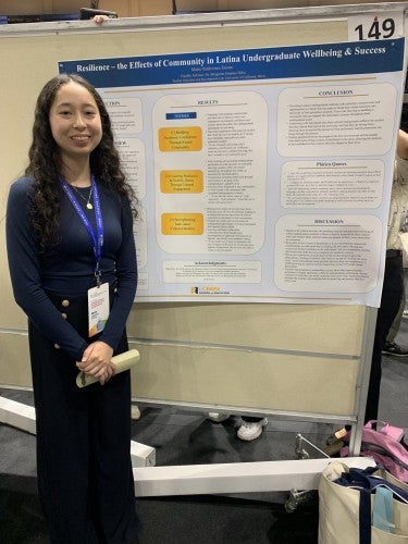 Maria Valdovinos poses for a photo in front of her poster that reads "Resilience – the Effects of Community in Latina Undergraduate Wellbeing & Success"