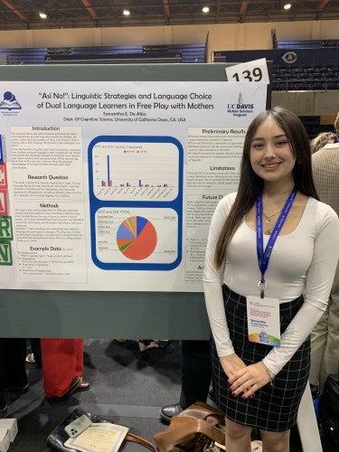 Samantha De Alba Sanchez poses for a photo in front of her poster that reads "'Asi No!': Linguistic Strategies and Language Choice of Dual Language Learners in Free Play with Mothers"