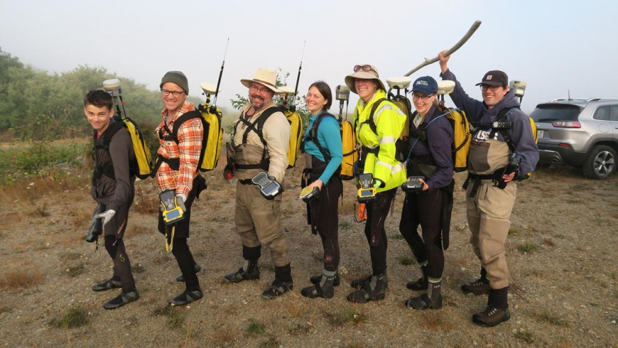 A group of seven people stand in a line, wearing GPS equipment, dressed for outdoor field work, and smiling.