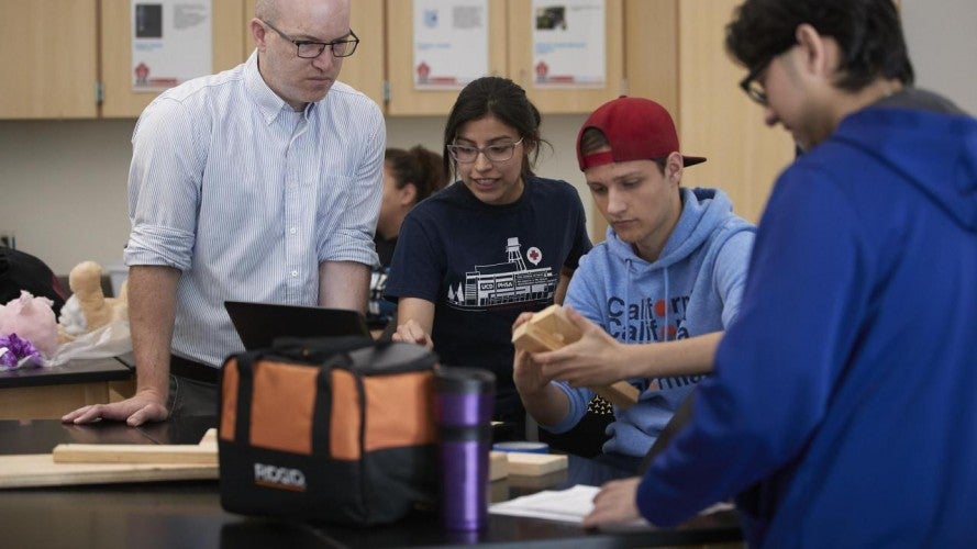 A man wearing a white button down and glasses looks at a project that three students are constructing with their hands.