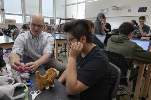 A man wearing a white button down and glasses reaches over and adjusts a project with his hands, while a student sits next to him and watches.
