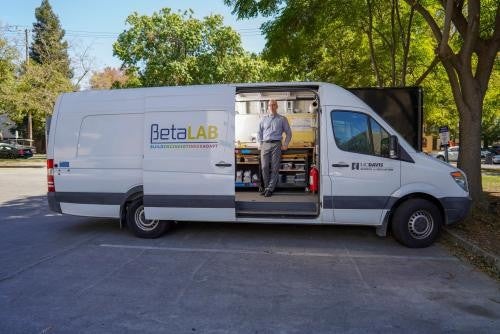 A white van with the words "Beta Lab" printed on the outside is parked in a parking lot. The van's side door is slid open, and inside a man wearing a blue button down and gray pants stands inside, posing for a camera and looking out the door.