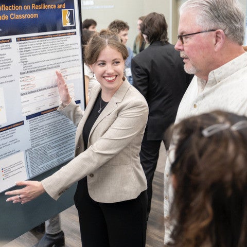 A woman wearing a blazer and black dress stands in front of a research poster and gestures toward it while smiling at a small group of people in front of her.