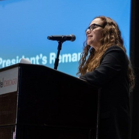 Michal Kurlaender stands at a podium and speaks into a microphone. She is backlit by a presentation screen that reads "AEFP President's Remarks."