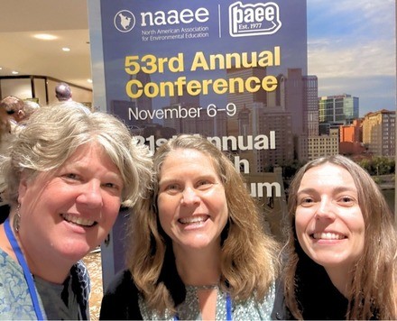 Three women pose together in front of a NAAEE banner.