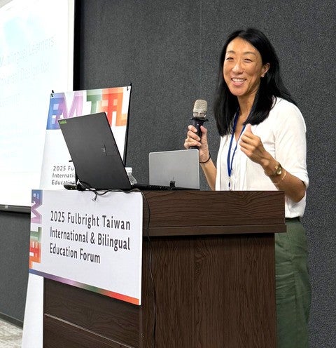 A woman wearing a white blouse and green pants stands at a podium and speaks into a microphone. Attached to the podium is a sign that reads "2025 Fulbright Taiwan International & Bilingual Education Forum."