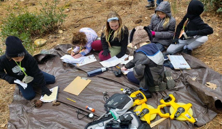A group of students sit on a tarp recording notes on paper