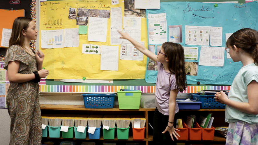 A young girl stands at the front of a classroom and points to a poster, while a teacher and other student standby watching.