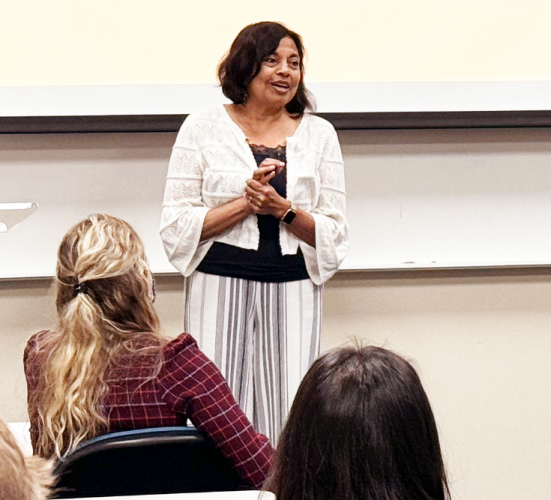 A woman stands in front of a projector screen and addresses an audience seated in front of her.