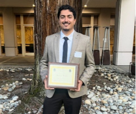 A man wearing a suit and tie stands in front of a tree and poses for a photo while holding a plaque in his hands.