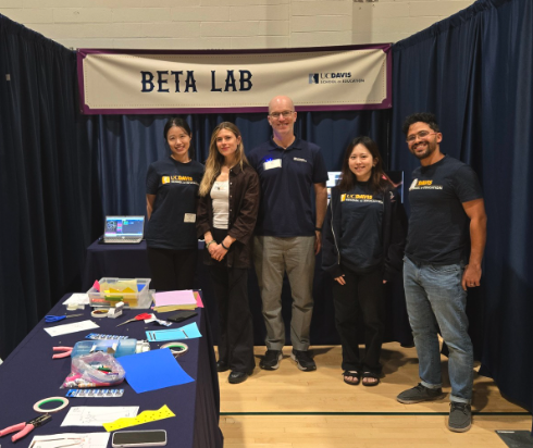 A group of five people stand in front of a sign that says "Beta Lab." In front of them is a table with construction paper, scissors, and other tools.