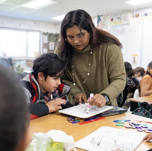 A woman wearing a green sweater leans over a desk where a young boy is sitting and writes on a handheld white board.