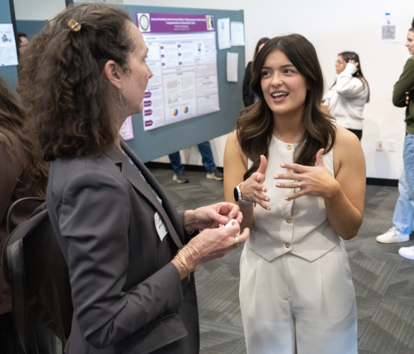 Two woman stand in front of a poster and speak to each other.