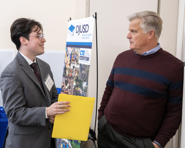 Two men speak to each other, standing in front of a poster advertising a school district.