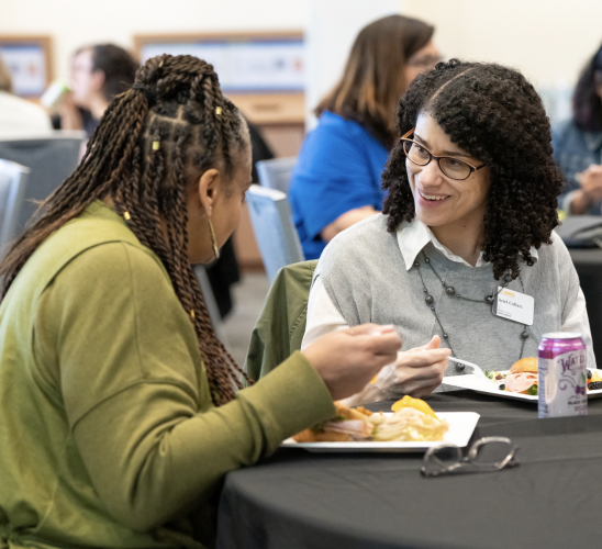 Two women, one wearing a green sweater and one wearing a grey sweater, sit together at a table and speak to each other while eating lunch.