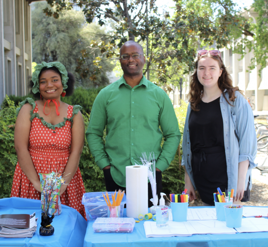 A man wearing a green shirt stands at a table with craft supplies. A woman wearing a red dress and a woman wearing a black dress and grey sweater stand on either side of him.