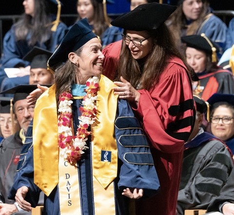 A faculty member wearing graduation regalia leans over a graduating PhD student to place a stoll on their shoulders.
