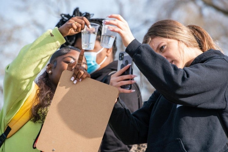Two female students hold clear plastic cups up to their faces and point to the juvenile fish inside them.