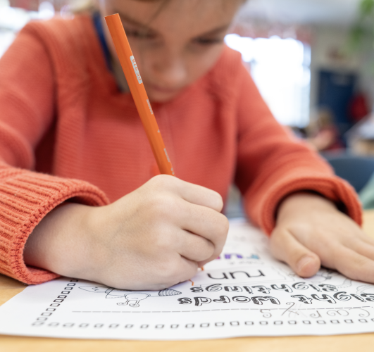 Close up of a young child seated at a classroom desk and writing with a pencil on a worksheet.